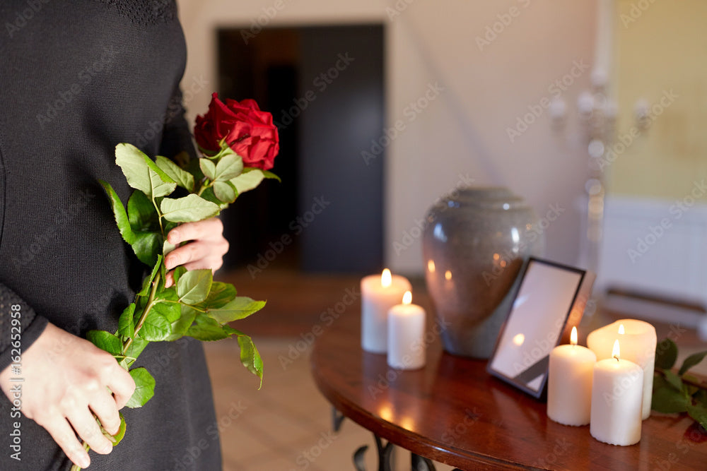 Metal cremation urn on table with candles, framed photo, and person holding red rose